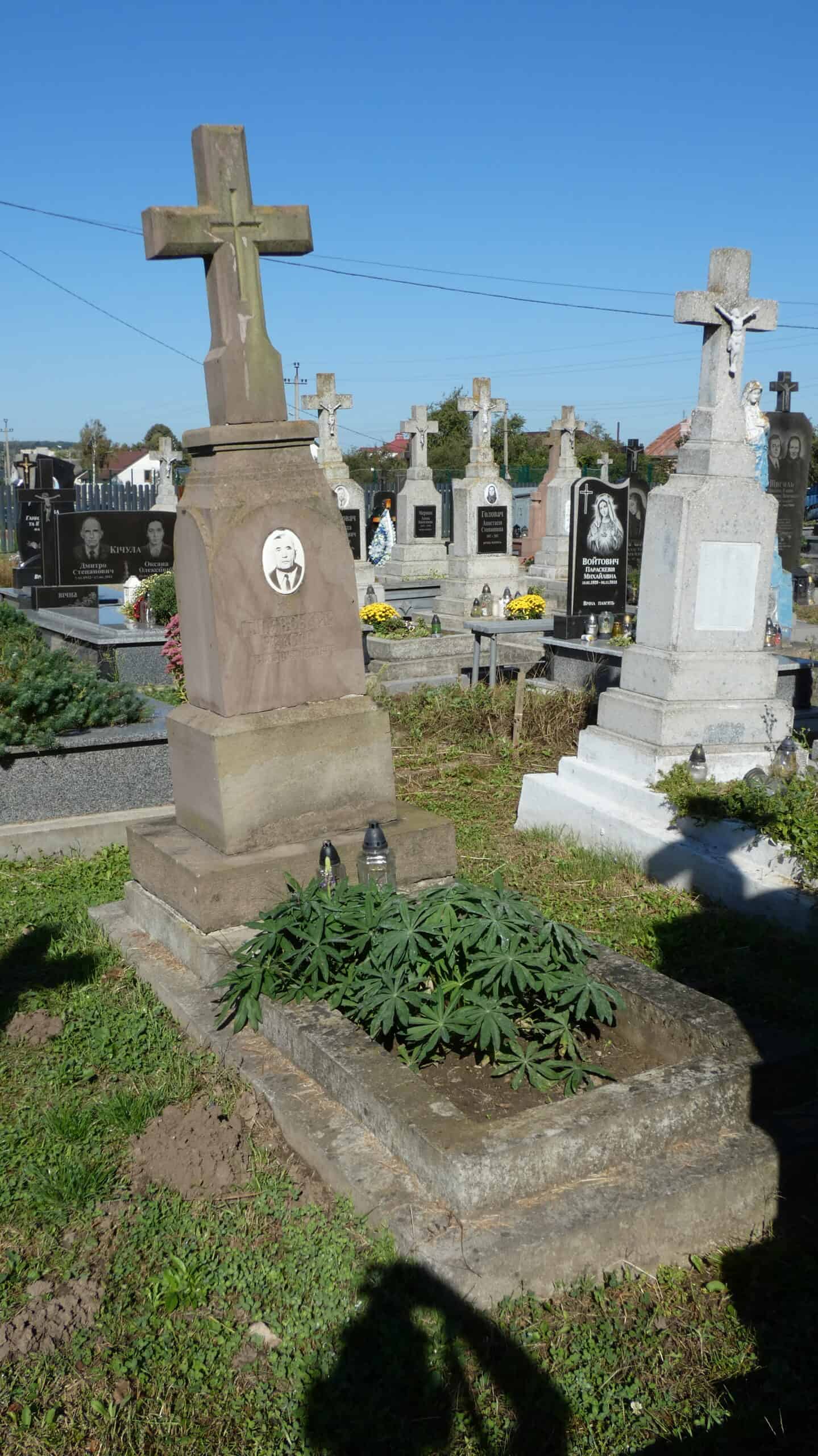 Меморіальний хрест на старому цвинтарі у Буцачі з покинутими могилами та квітами.