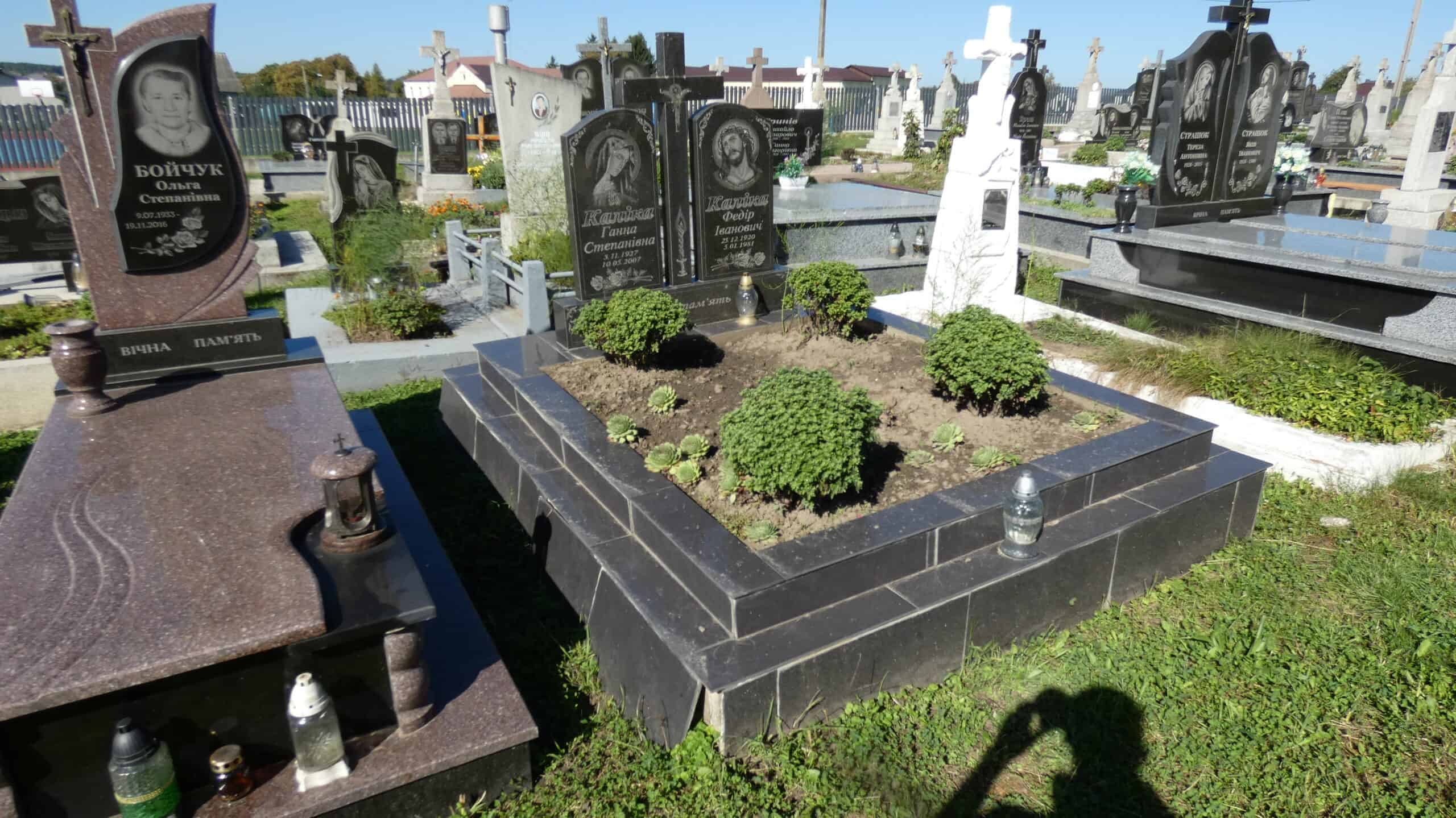 Могила з чорними пам’ятниками та зеленими кущами на цвинтарі Бучач.