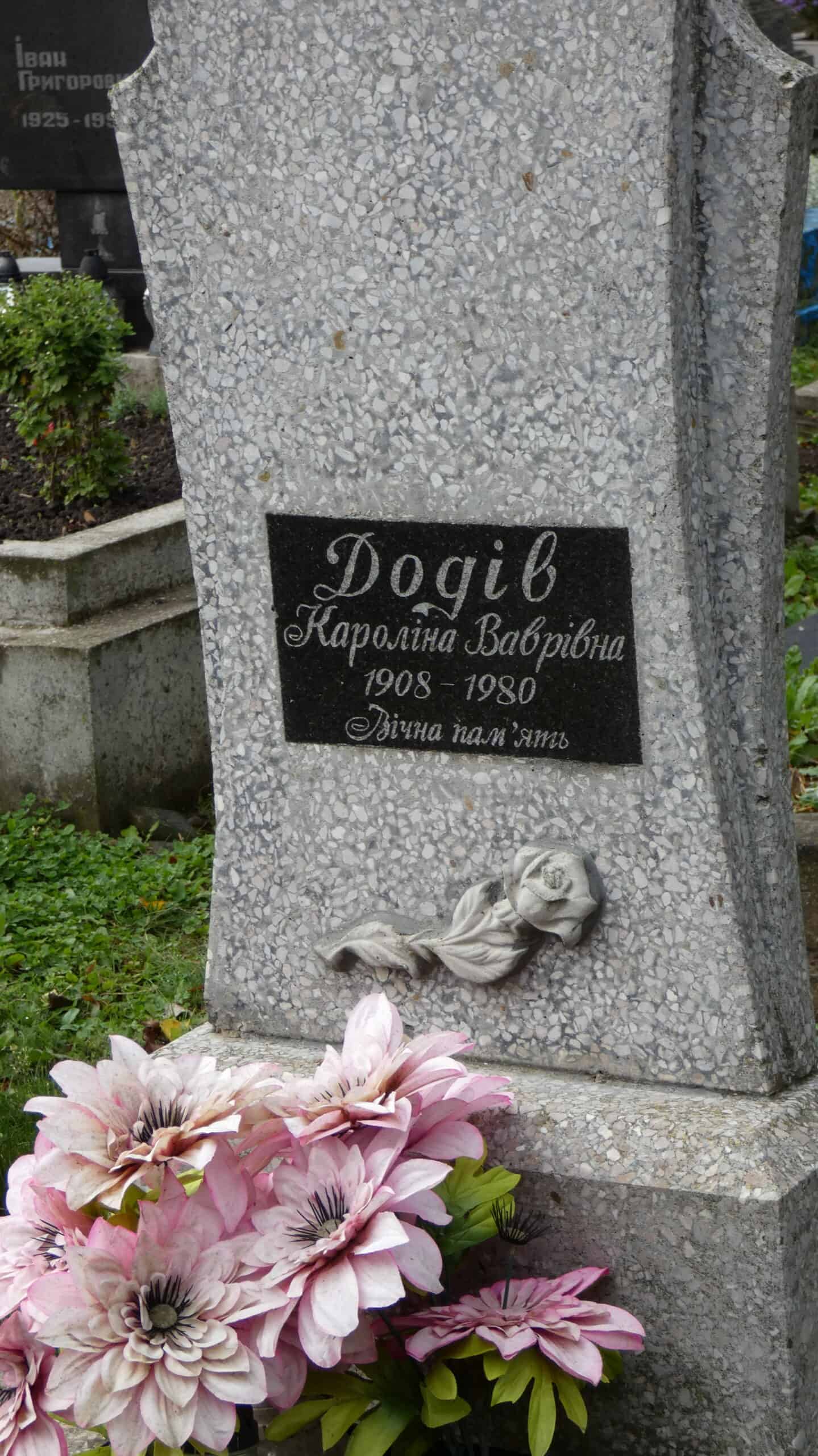 Квіти на могилі, меморіальний пам'ятник у цвинтарі Бучач. Хрест з ім'ям та датами, поруч квіти для поминання.