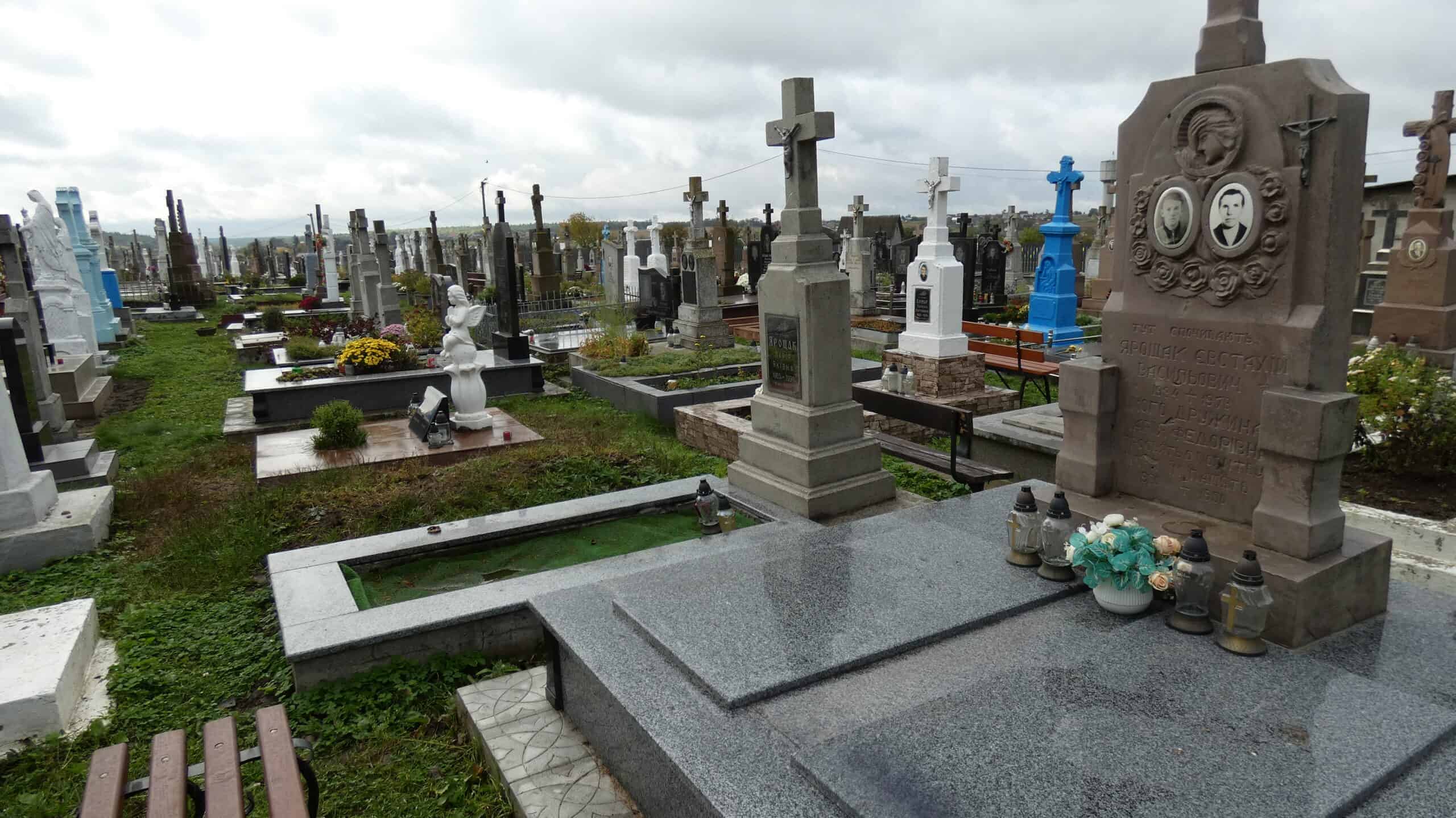 Могила з пам'ятником із хрестом та фотографіями родини, цвинтар у Буцьках, велика кількість поховань, квіти та лампадки.