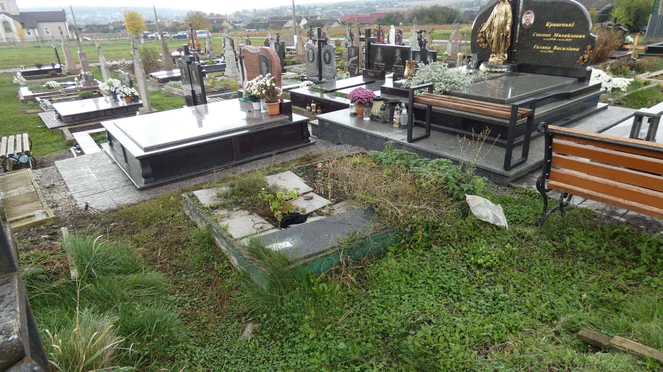 Білий та чорний надмогильний пам'ятник на цвинтарі у Буцачі, з квітами та лампадками, огороджений металевим парканом.