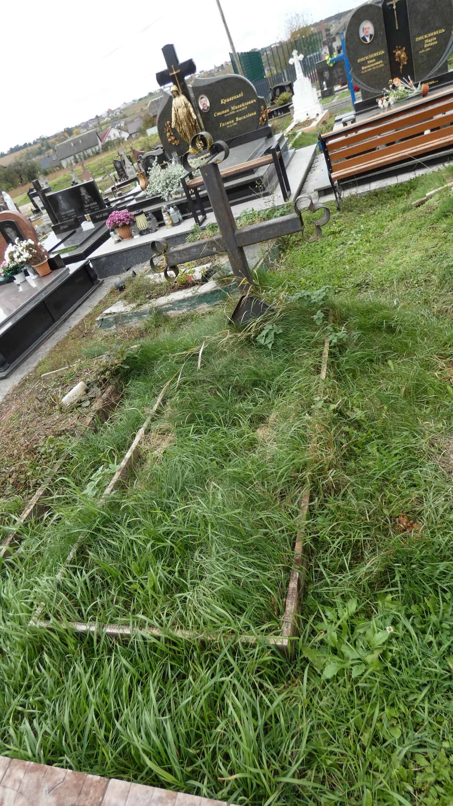 Зламаний хрест на цвинтарі, покреслений і пошкоджений, з ознаками руйнування та зростаючою травою навколо.